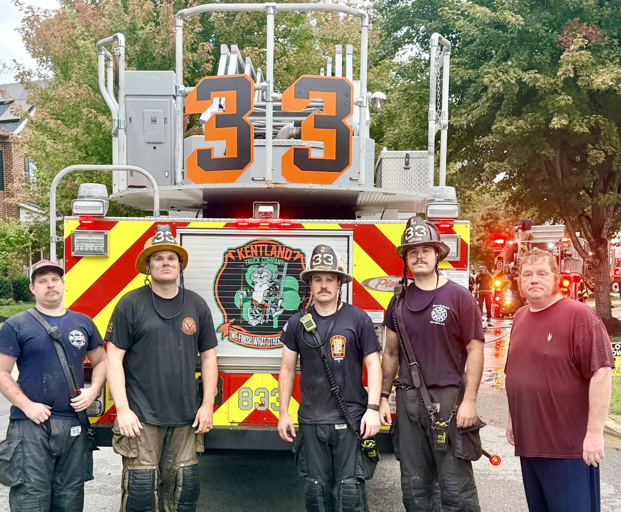 Four firefighters in uniform and one man in civilian clothes standing in front of a Kentland Truck Company fire engine labeled with the number 33.