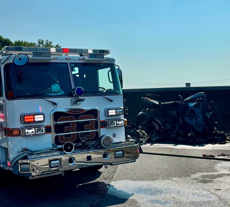 A fire truck is parked near the wreckage of a severely damaged vehicle on a sunny day, with debris scattered across the road.