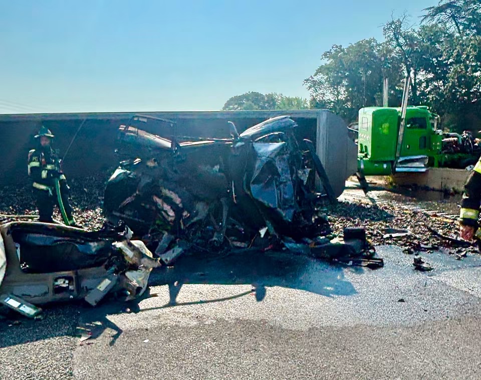 Two firefighters stand near the wreckage of a crushed vehicle and an overturned truck on a road, with debris scattered and a green semi-truck cab in the background.