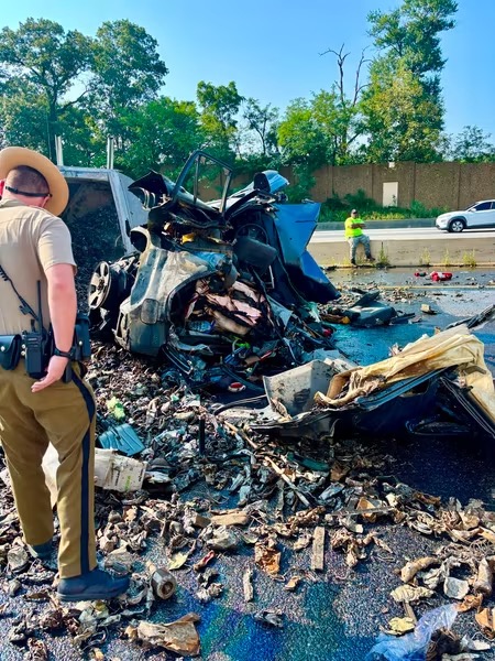 A police officer stands near the wreckage of a severely damaged vehicle and debris scattered across a road, with another worker in the background and trees lining the scene.