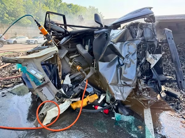 A severely wrecked car with crumpled metal and rescue equipment attached, sitting in a parking lot. The interior and engine are exposed, and debris surrounds the vehicle.