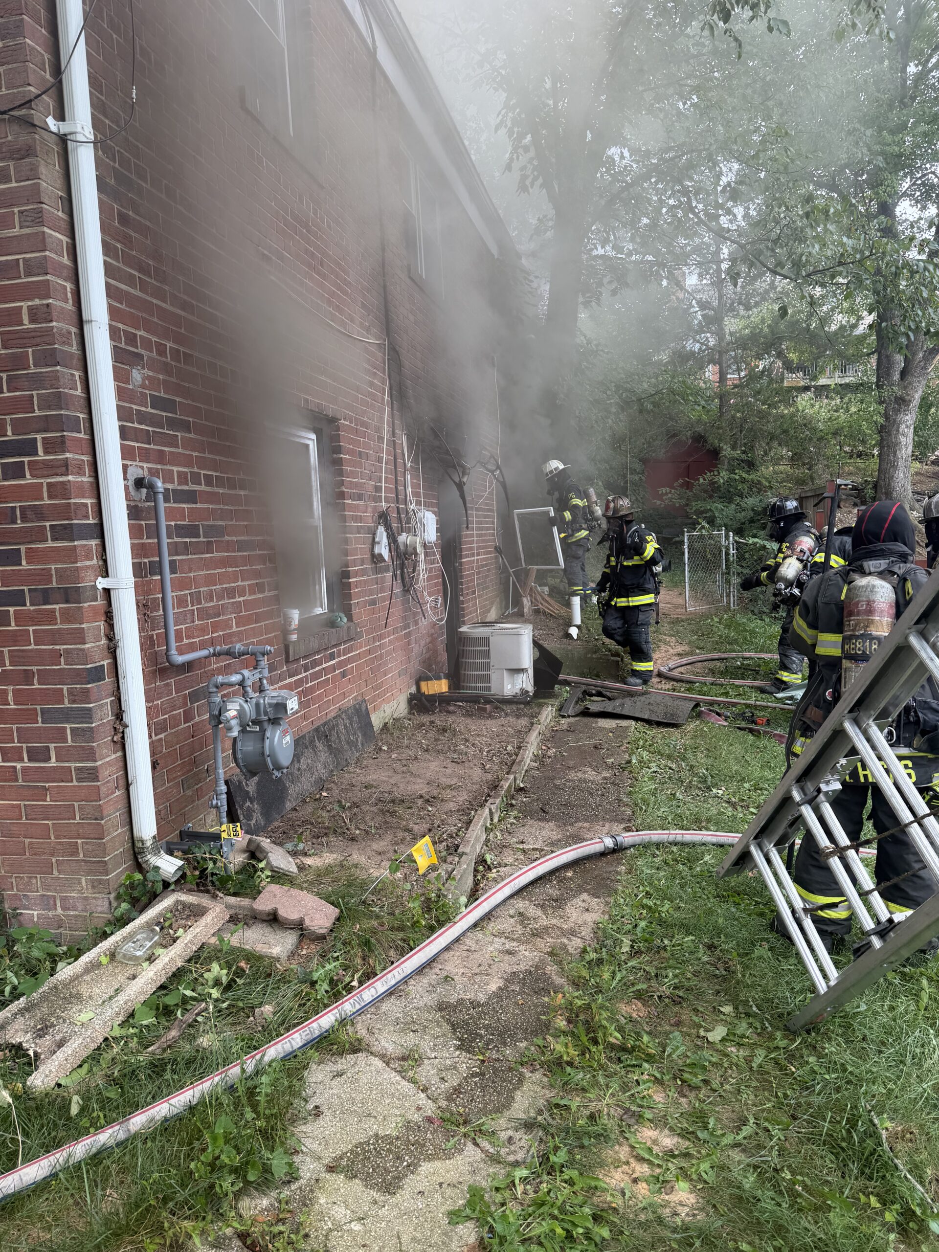 Firefighters in full gear work outside a brick building with smoke pouring from a window. Ladders, hoses, and equipment are scattered on the ground near the scene.