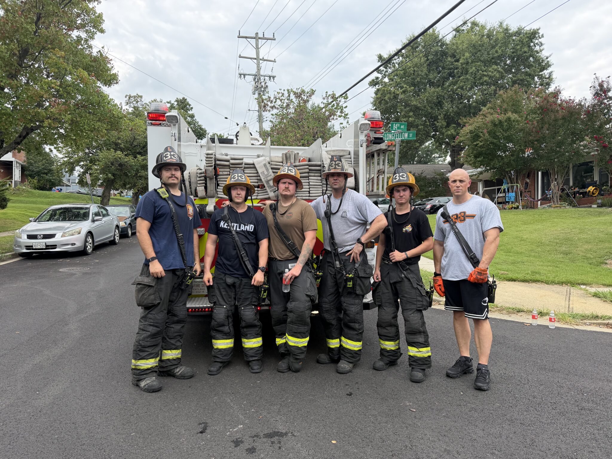 Six firefighters in gear stand together in front of a fire truck on a suburban street. Trees, power lines, and houses are visible in the background.