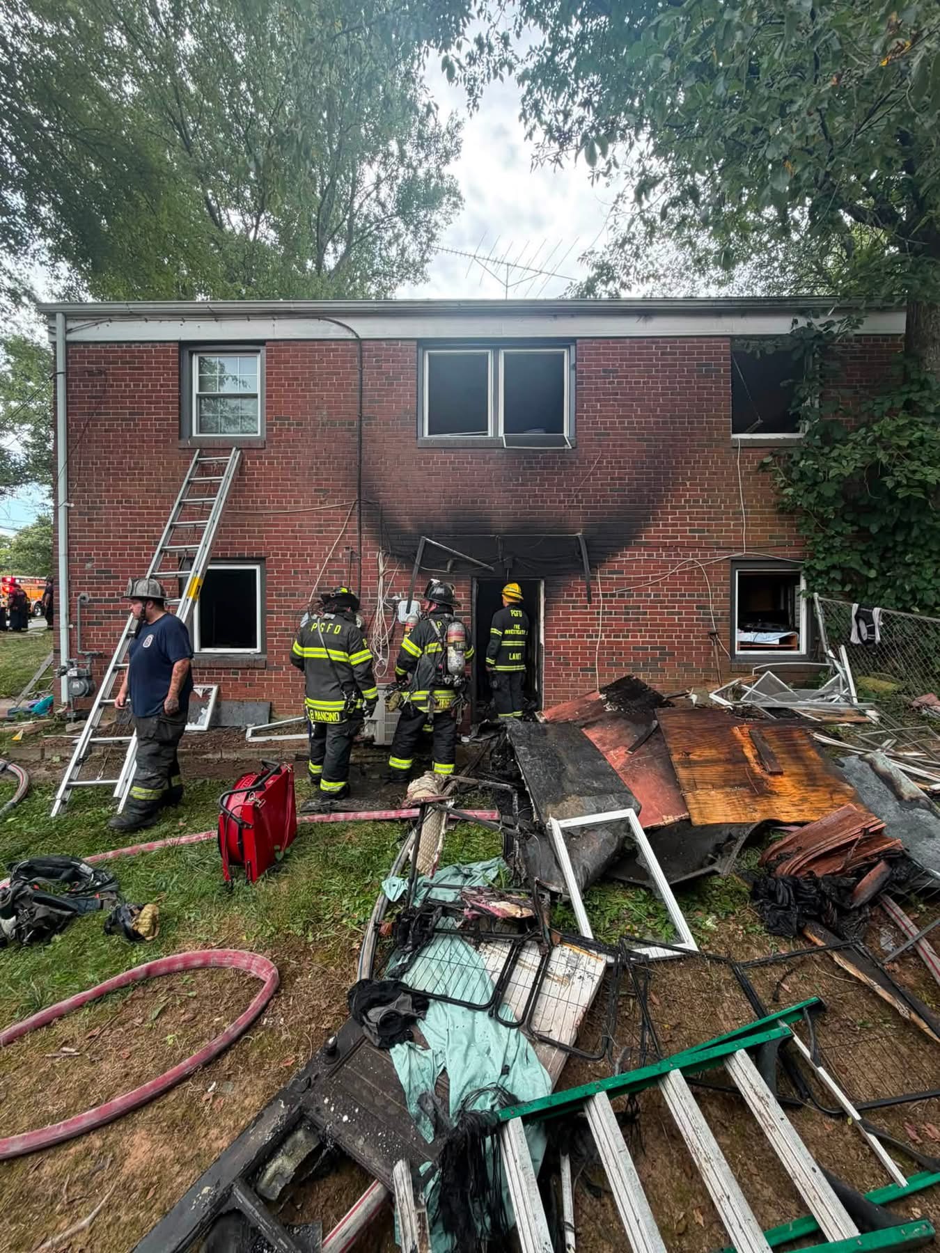 Firefighters in gear stand in front of a brick building with visible fire damage around a blackened doorway. Debris, ladders, and fire hoses are scattered across the lawn in the foreground.