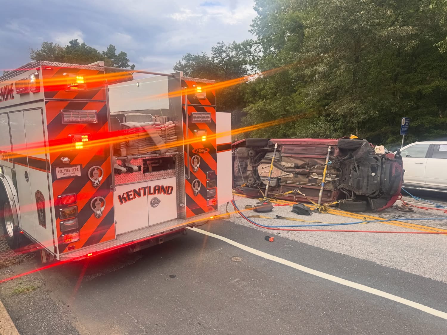 A fire truck with flashing lights is parked near an overturned car on its side. Emergency equipment is visible, and the scene appears to be an accident on a road with trees in the background.