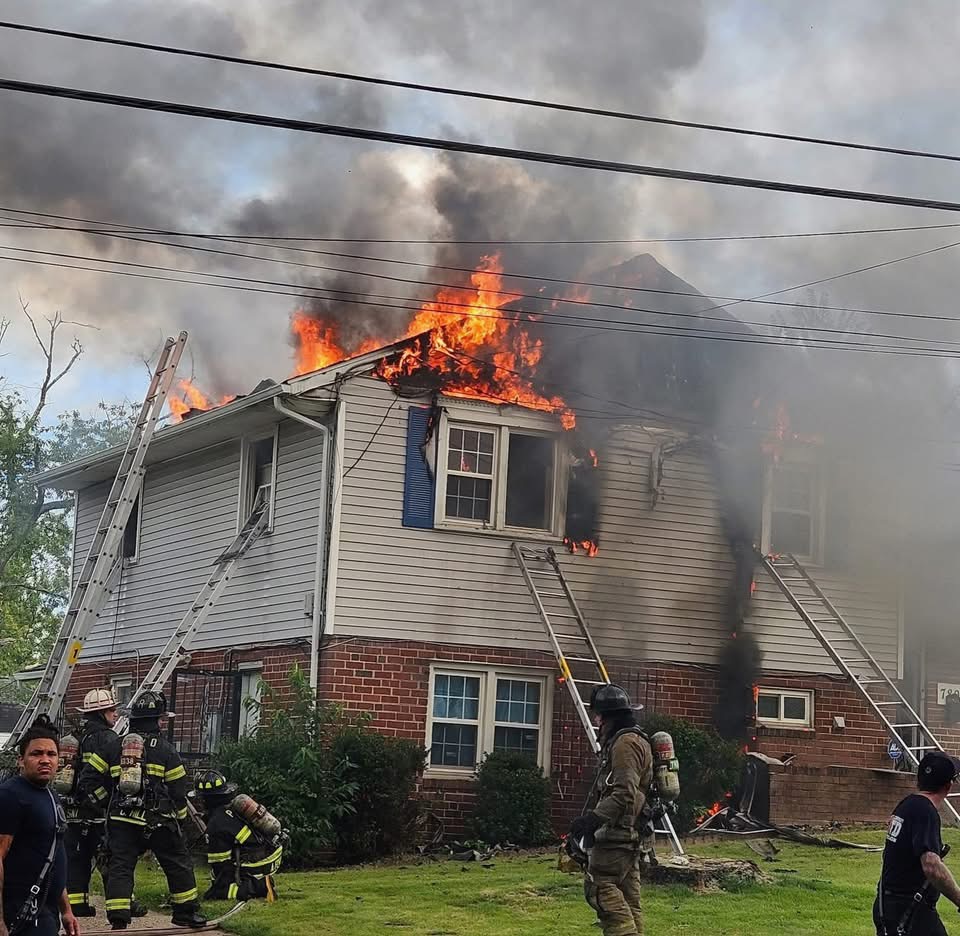 A house is on fire with flames and smoke coming from the roof. Firefighters and ladders surround the building as they work to control the blaze.