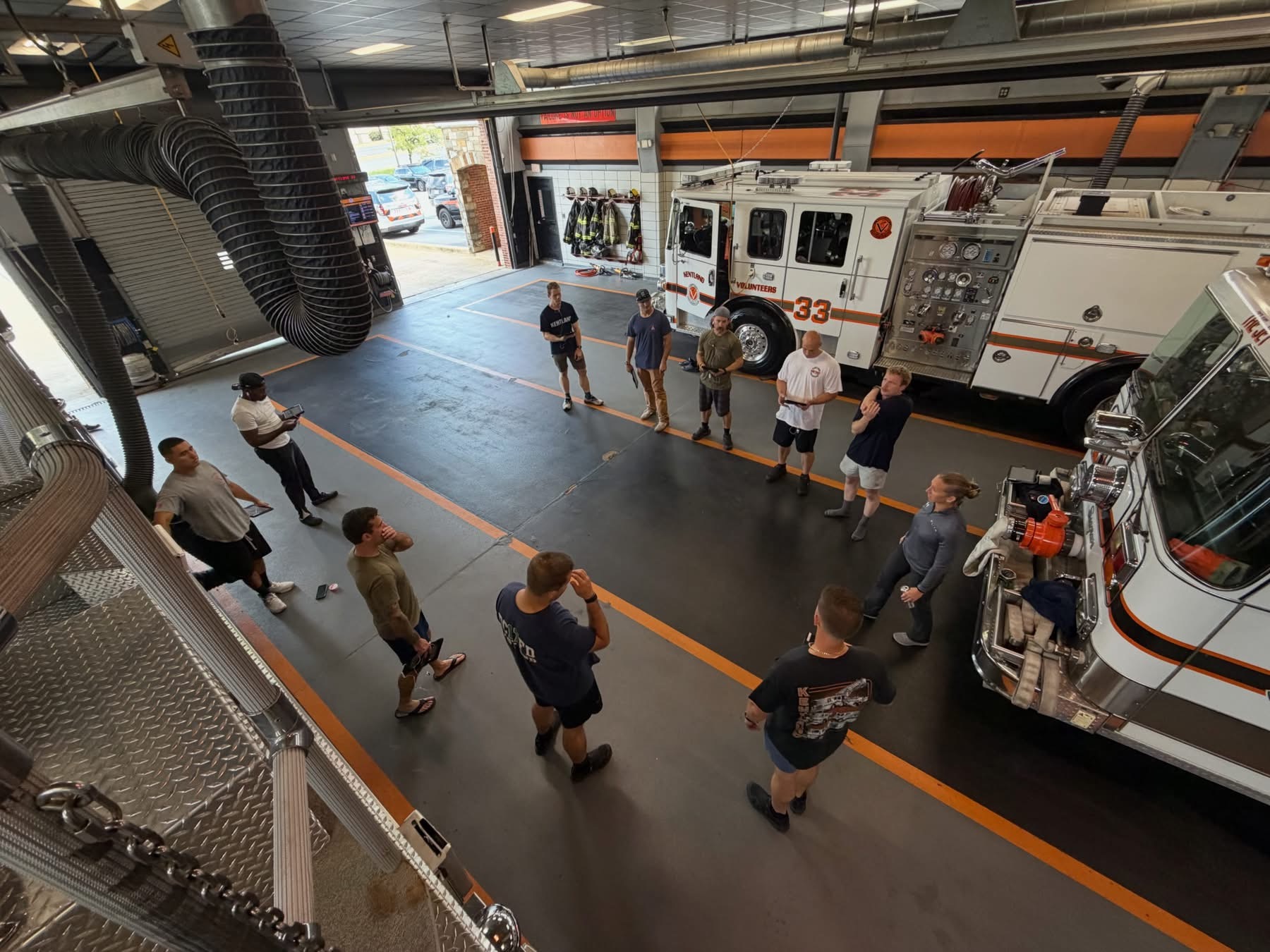A group of people, some in uniforms, stand in a fire station garage near two fire trucks, having a discussion. Firefighting gear hangs on the wall in the background.