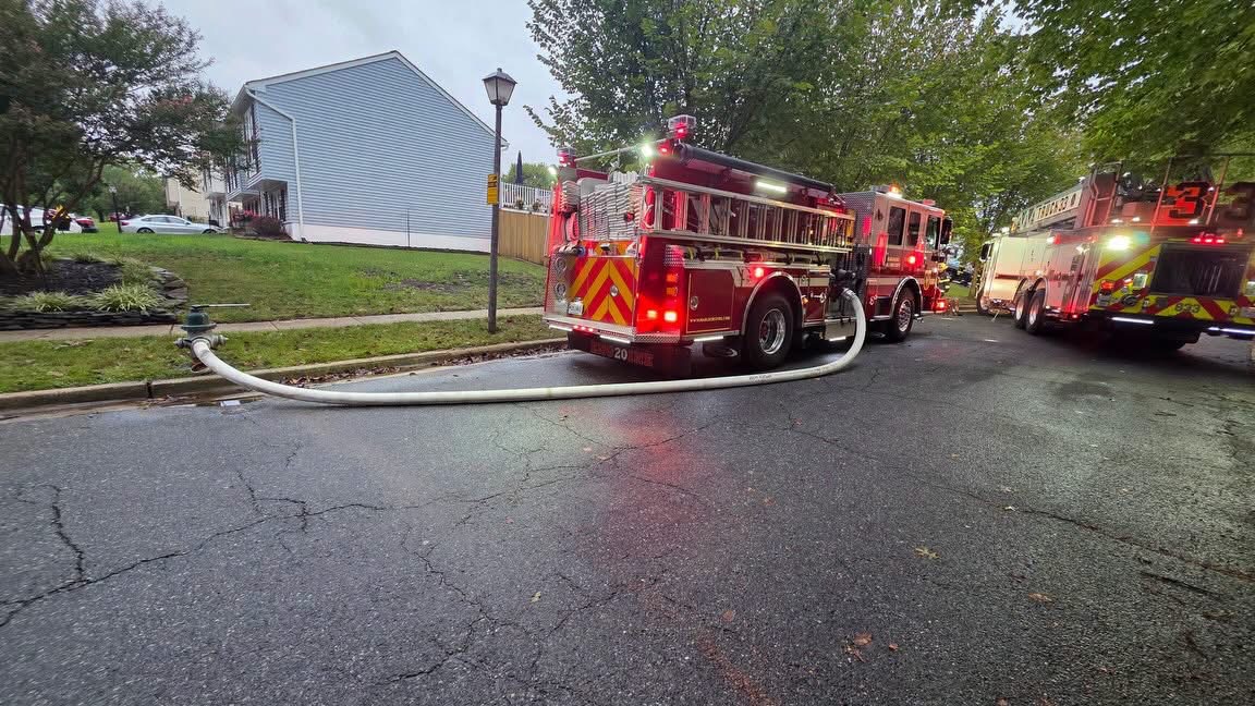 Two red fire trucks parked on a residential street, with a fire hose connected to a hydrant in the foreground.
