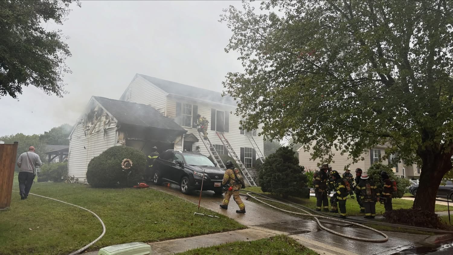 Firefighters in turnout gear work to extinguish a fire at a two-story residential house as smoke pours from the roof and a car sits parked in the driveway.