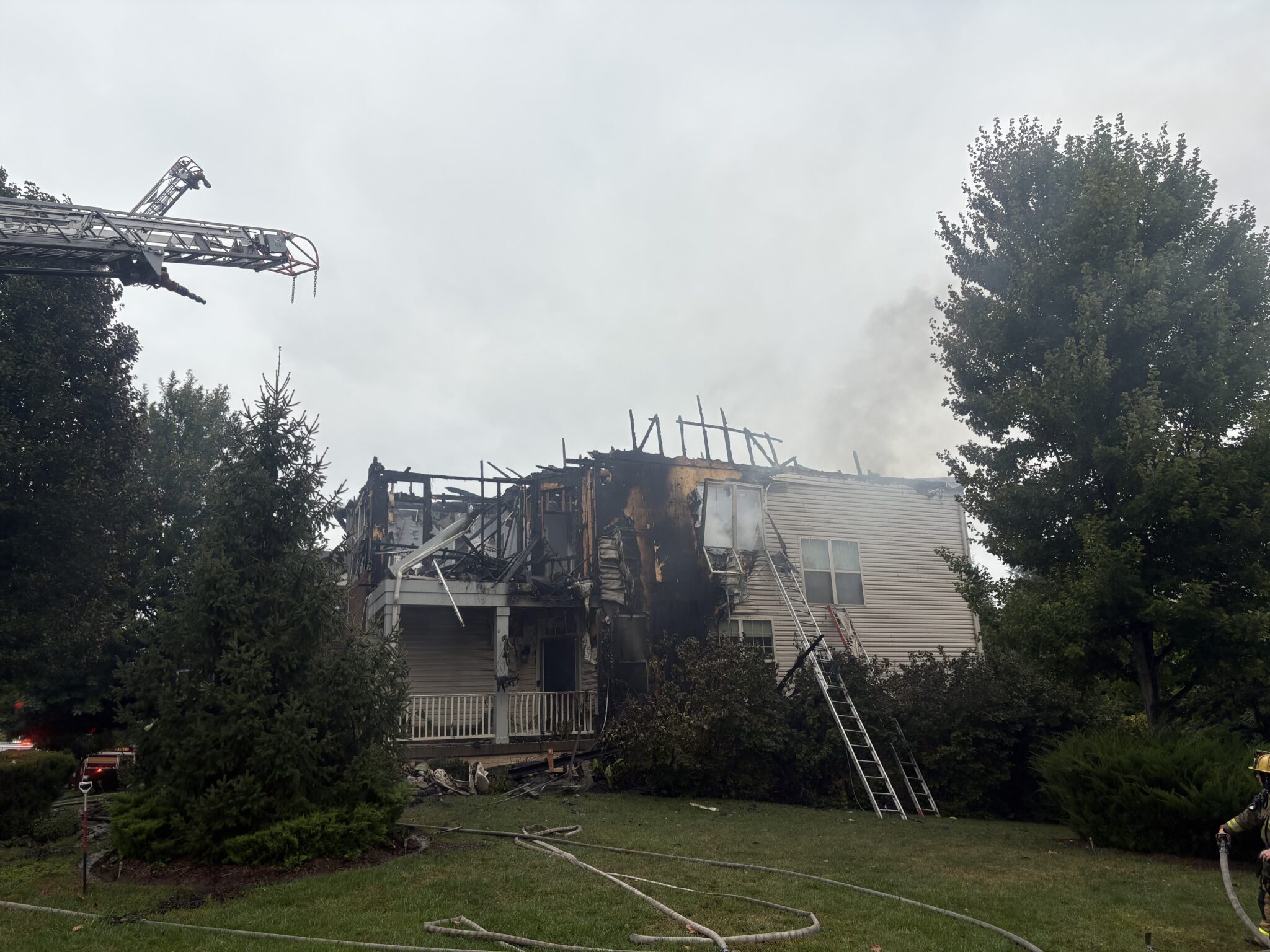 A house with severe fire damage to its roof and siding, with a fire department ladder truck and a firefighter visible on the lawn.