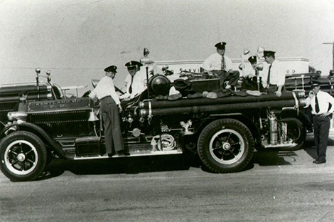 Black-and-white photo of five uniformed firefighters standing and sitting on an old-fashioned fire truck, equipped with hoses and equipment, parked on a street.