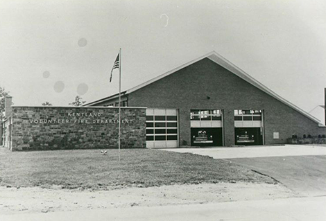 Black and white photo of the Kentland Volunteer Fire Department building, featuring a large triangular roof, an American flag on a pole, and two garage doors with emergency vehicles inside.