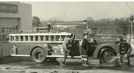 Black-and-white photo of a vintage fire truck parked outside a fire department building, with several firefighters standing next to the truck. The truck has ladders and hoses attached.